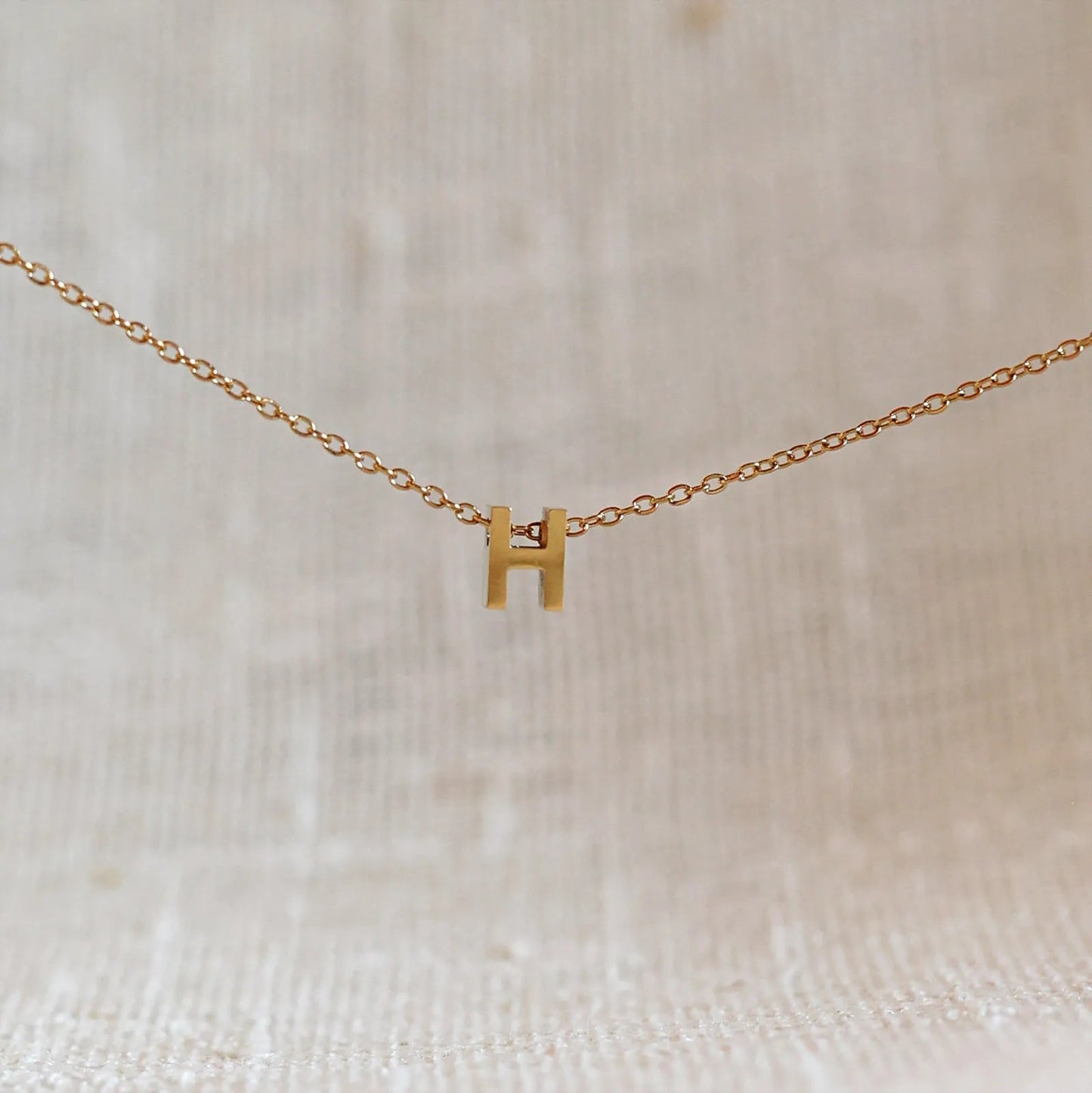 Collier L'instant avec pendentif lettre H doré en acier inoxydable, suspendu au centre d'une maille fine sur fond beige.