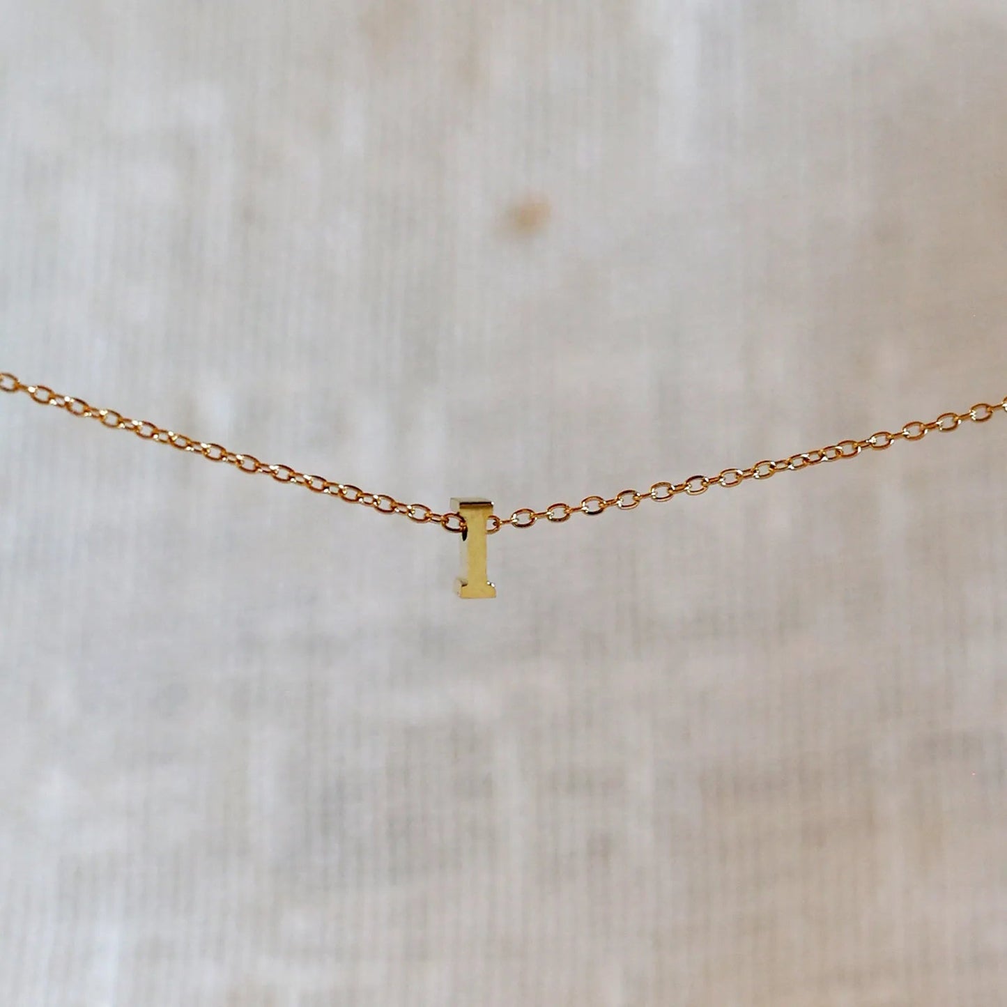 Collier L'instant avec pendentif lettre I doré en acier inoxydable, suspendu au centre d'une maille fine sur fond beige.