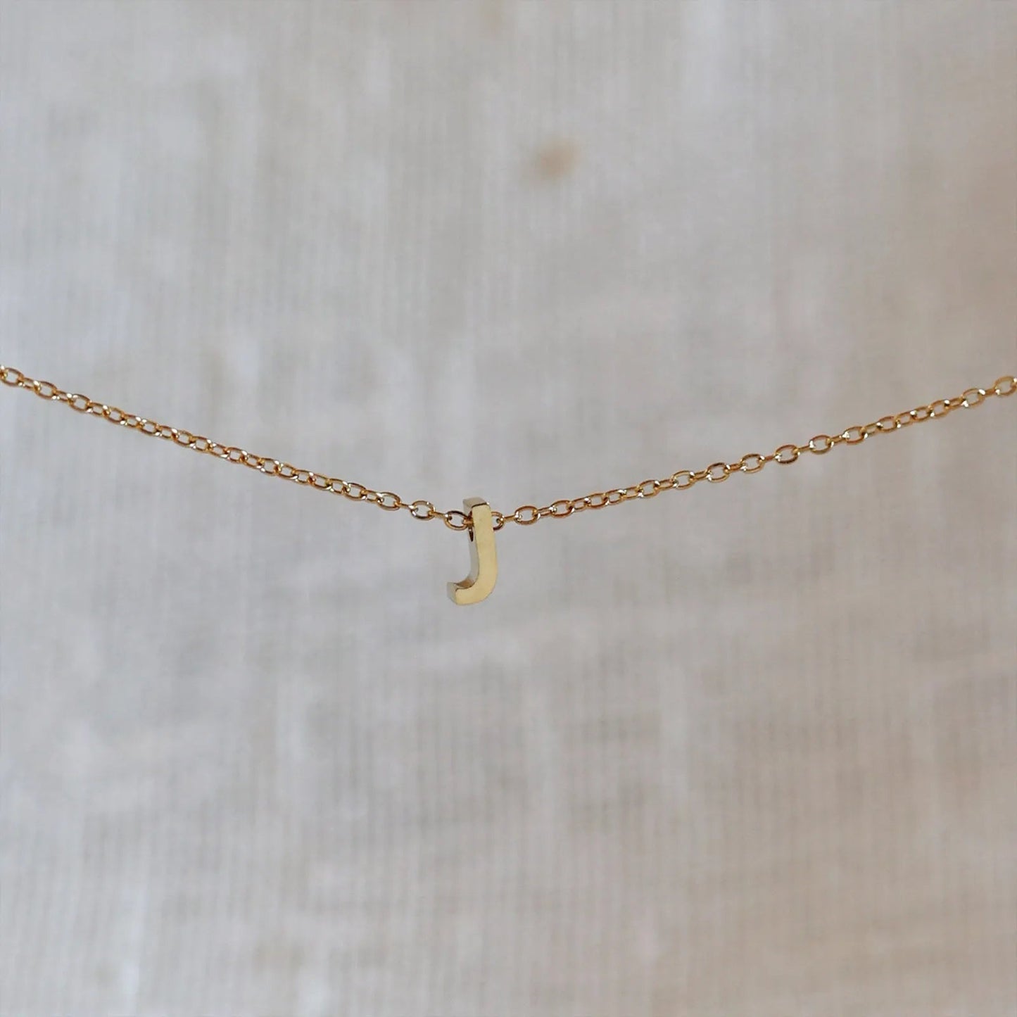 Collier L'instant avec pendentif lettre J doré en acier inoxydable, suspendu au centre d'une maille fine sur fond beige.
