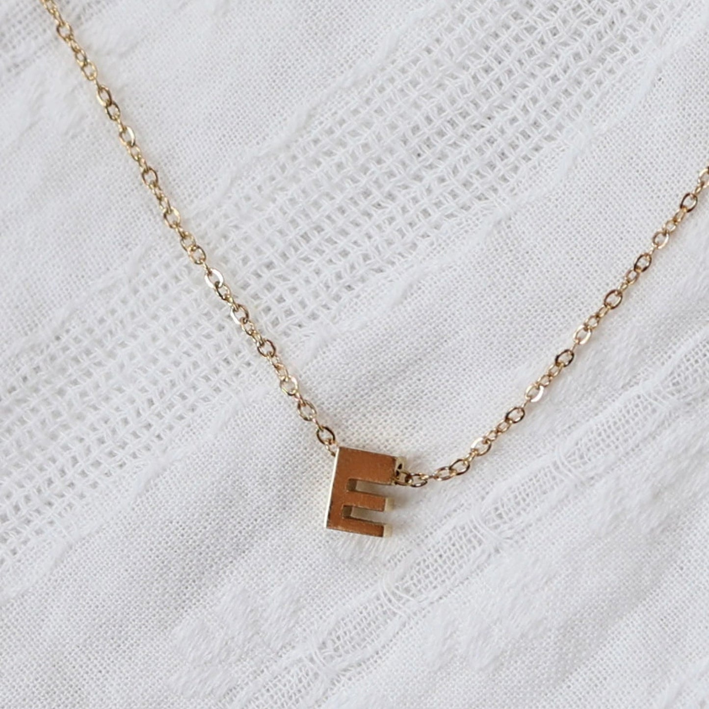 Zoom sur le collier L'instant, pendentif lettre E en acier inoxydable doré, capturé sur tissu beige texturé.