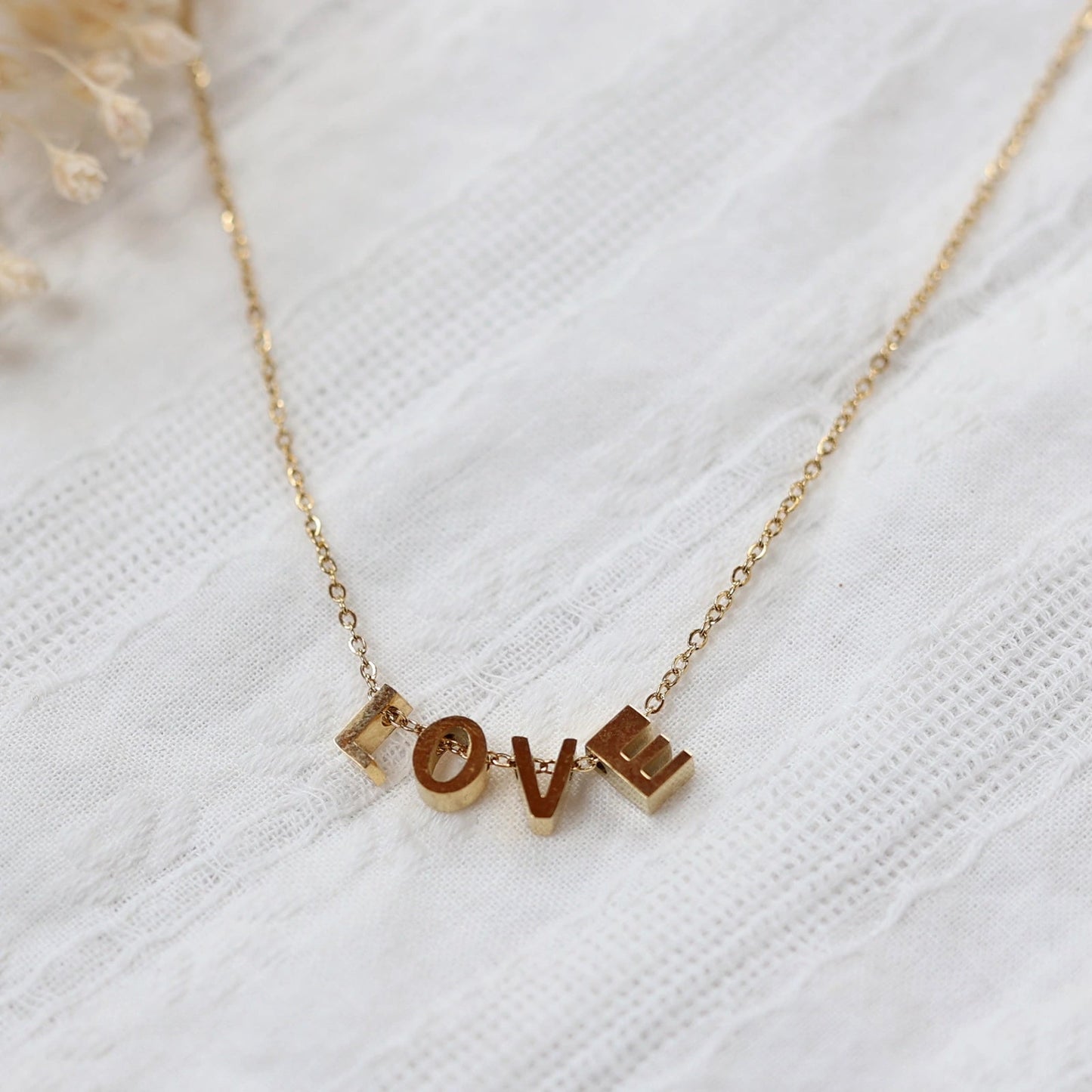 Collier L'instant avec pendentifs lettres LOVE en acier inoxydable doré, bijou délicat posé sur un tissu beige texturé.