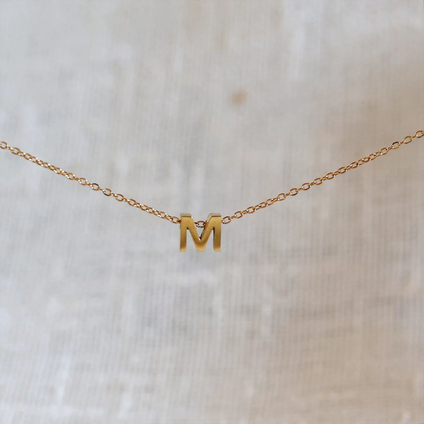 Collier L'instant avec pendentif lettre M doré en acier inoxydable, suspendu au centre d'une maille fine sur fond beige.