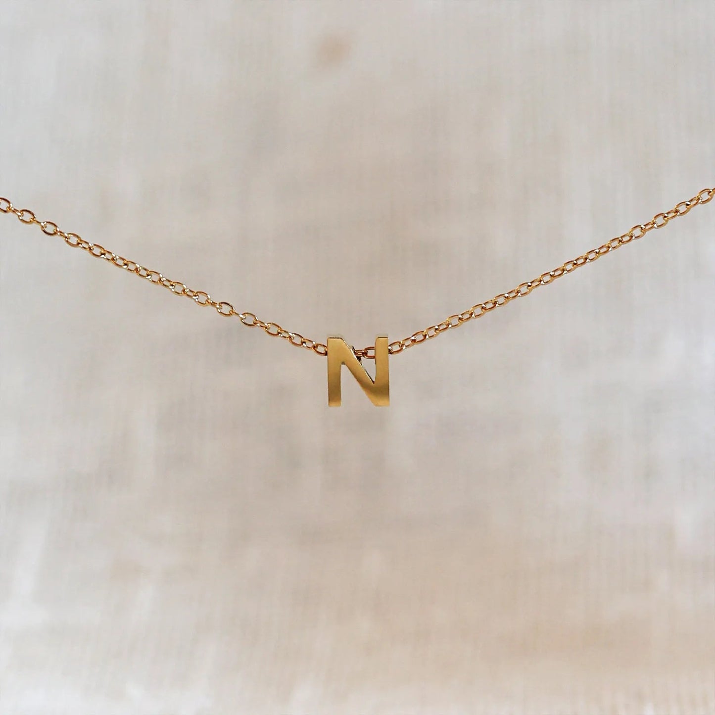 Collier L'instant avec pendentif lettre N doré en acier inoxydable, suspendu au centre d'une maille fine sur fond beige.