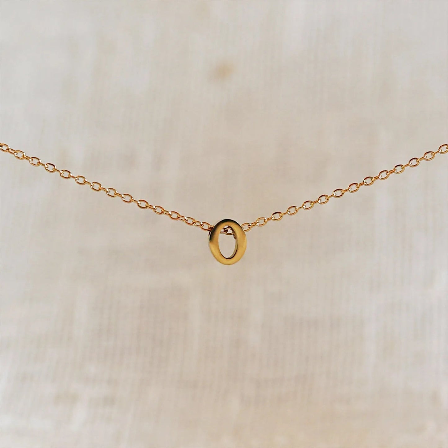 Collier L'instant avec pendentif lettre O doré en acier inoxydable, suspendu au centre d'une maille fine sur fond beige.
