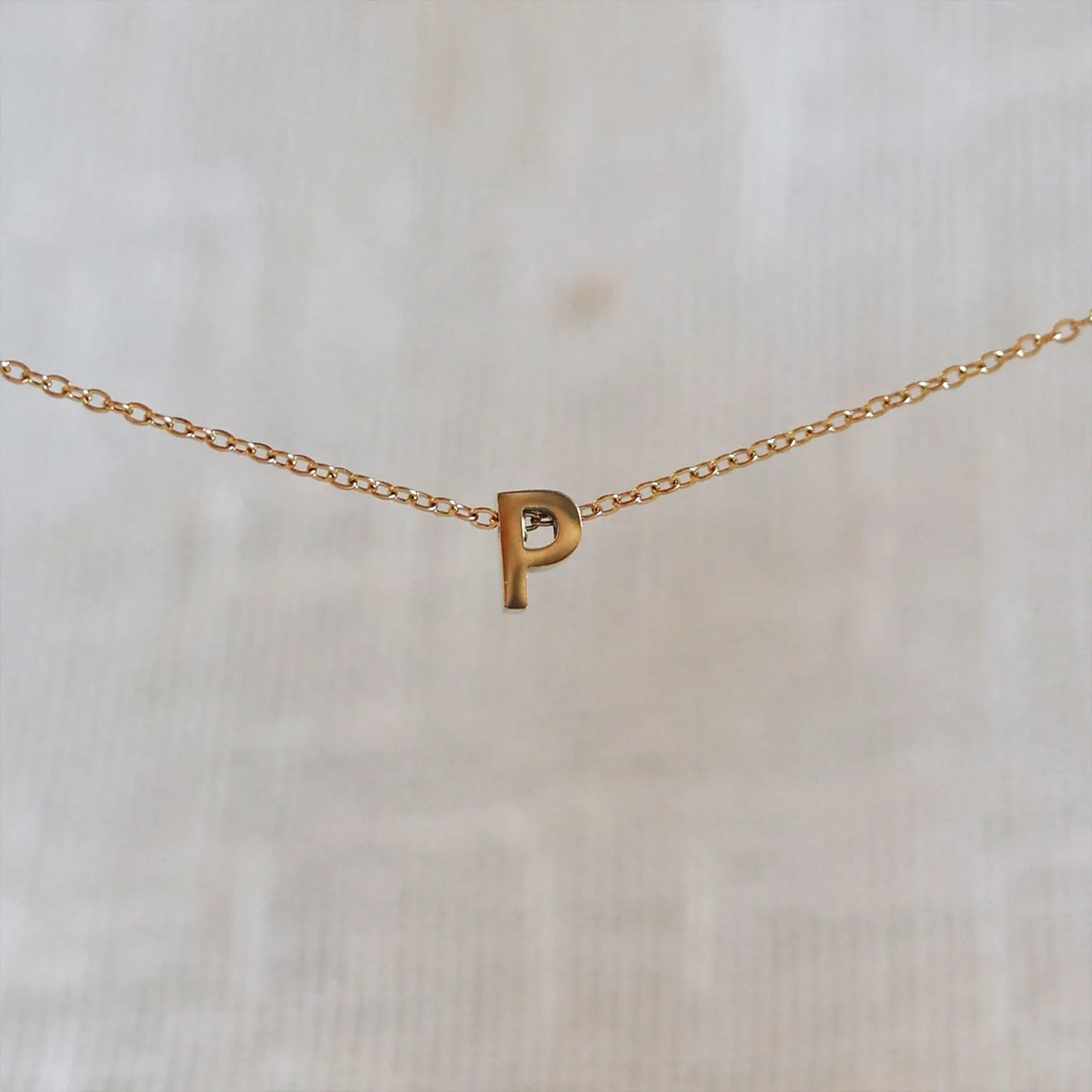 Collier L'instant avec pendentif lettre P doré en acier inoxydable, suspendu au centre d'une maille fine sur fond beige.
