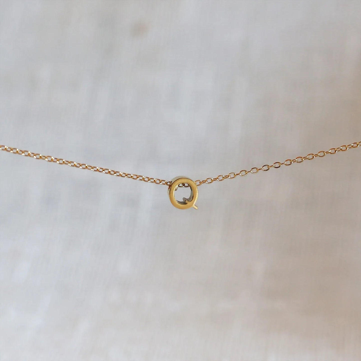 Collier L'instant avec pendentif lettre Q doré en acier inoxydable, suspendu au centre d'une maille fine sur fond beige.