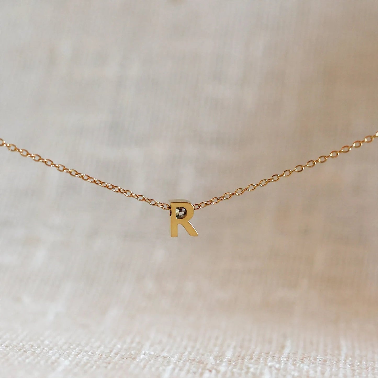 Collier L'instant avec pendentif lettre R doré en acier inoxydable, suspendu au centre d'une maille fine sur fond beige.