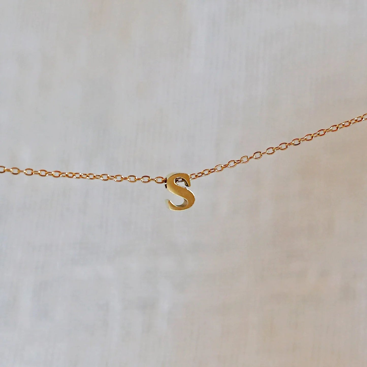 Collier L'instant avec pendentif lettre S doré en acier inoxydable, suspendu au centre d'une maille fine sur fond beige.