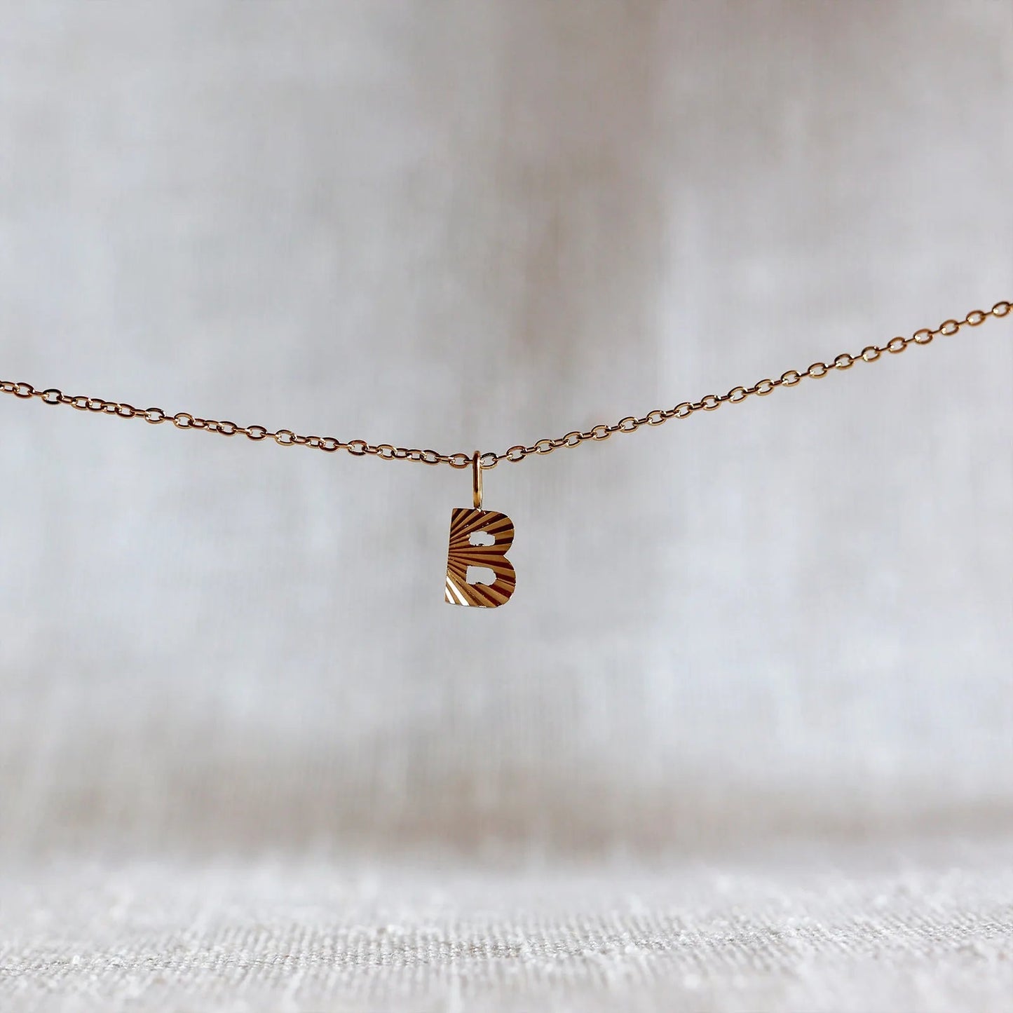 Collier "Je suis" avec pendentif lettre B striée en acier doré, suspendu sur un fond en tissu beige texturé.