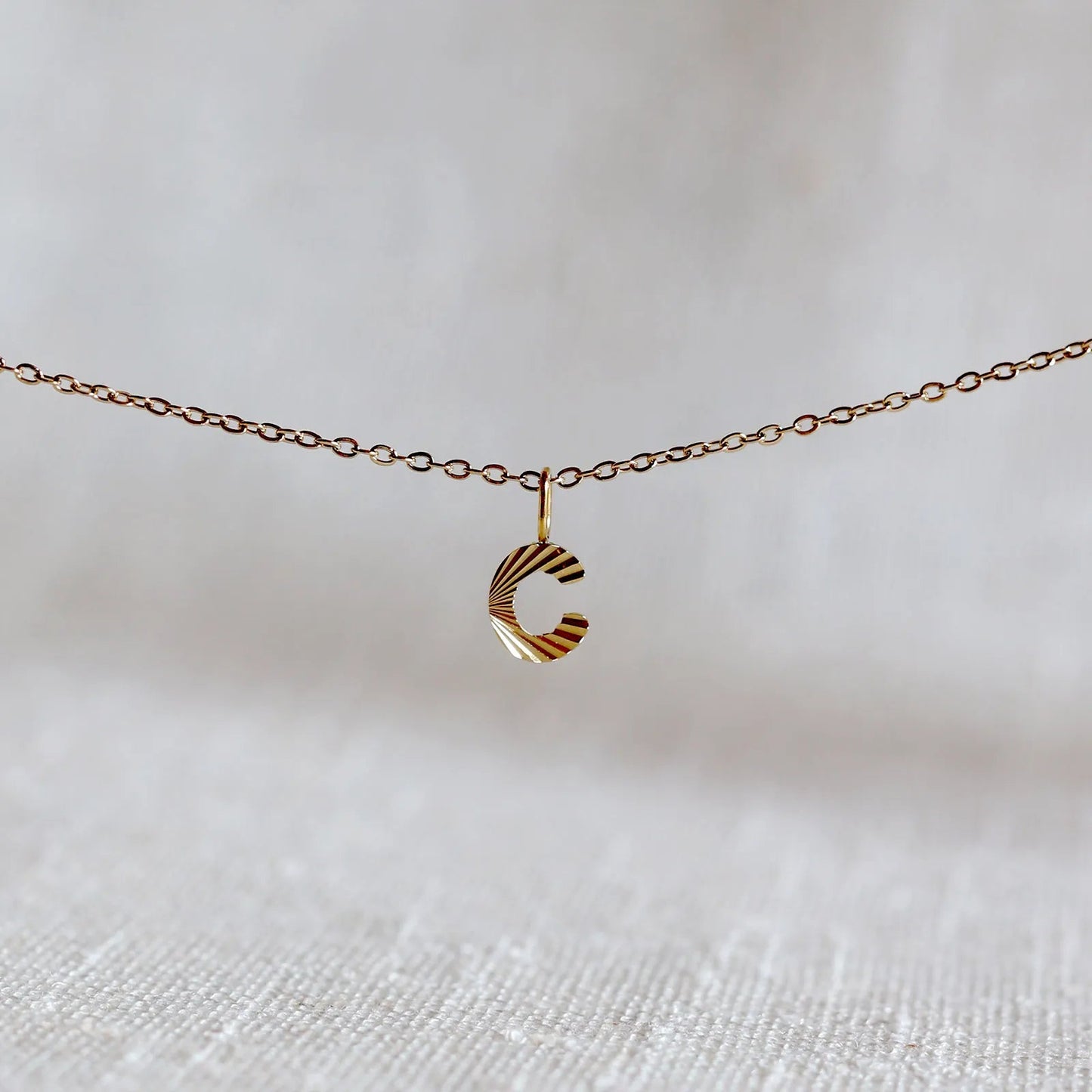 Collier "Je suis" avec pendentif lettre C striée en acier doré, suspendu sur un fond en tissu beige texturé.