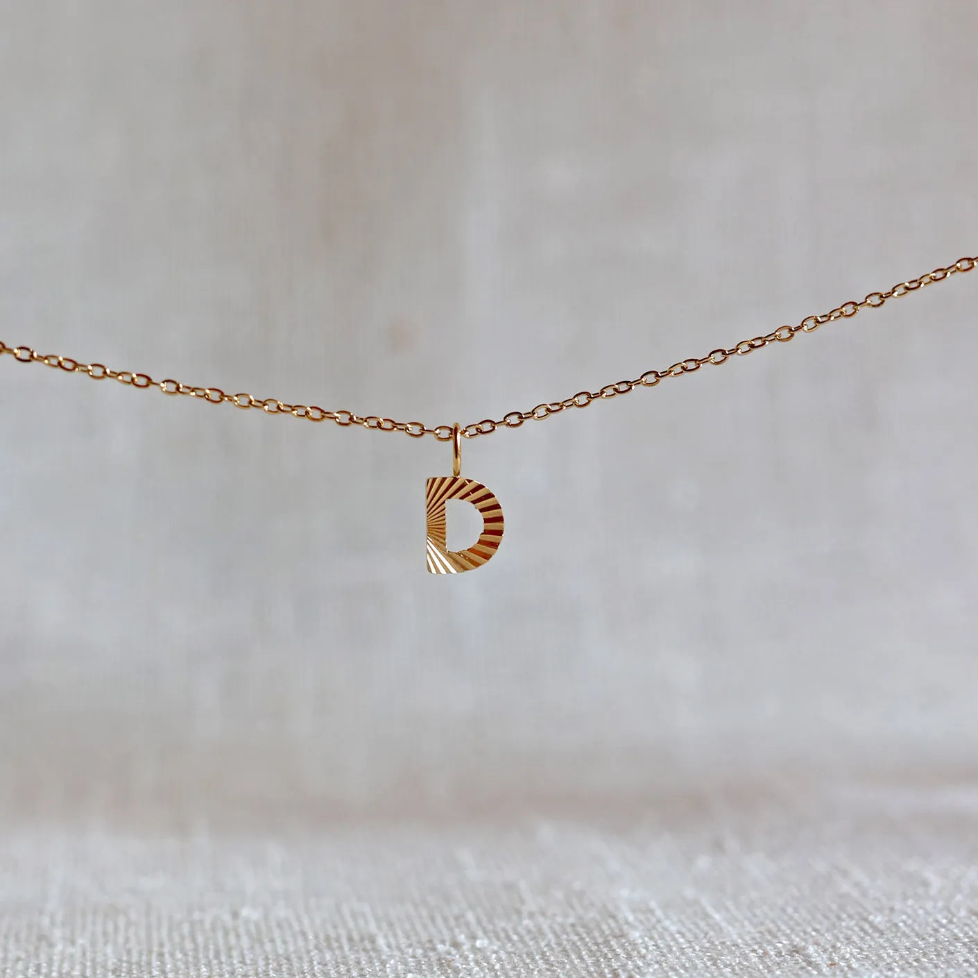 Collier "Je suis" avec pendentif lettre D striée en acier doré, suspendu sur un fond en tissu beige texturé.