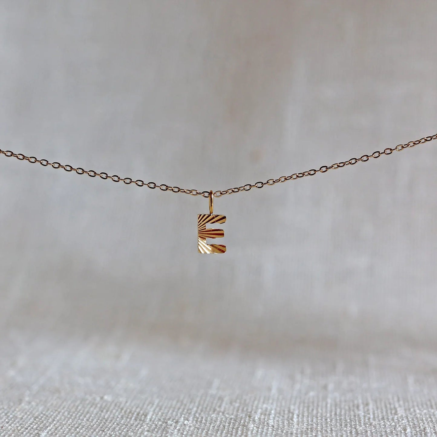 Collier "Je suis" avec pendentif lettre E striée en acier doré, suspendu sur un fond en tissu beige texturé.