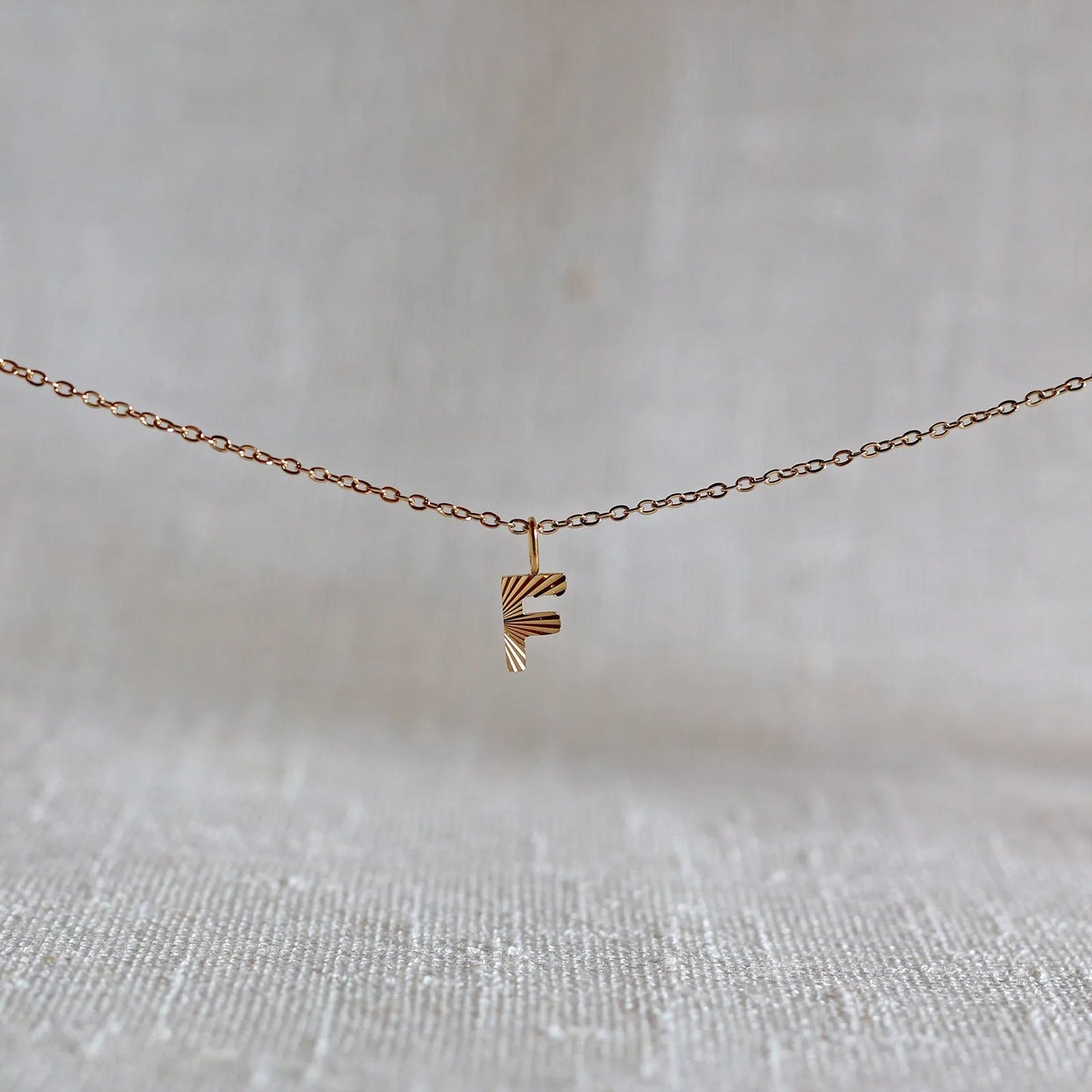 Collier "Je suis" avec pendentif lettre F striée en acier doré, suspendu sur un fond en tissu beige texturé.