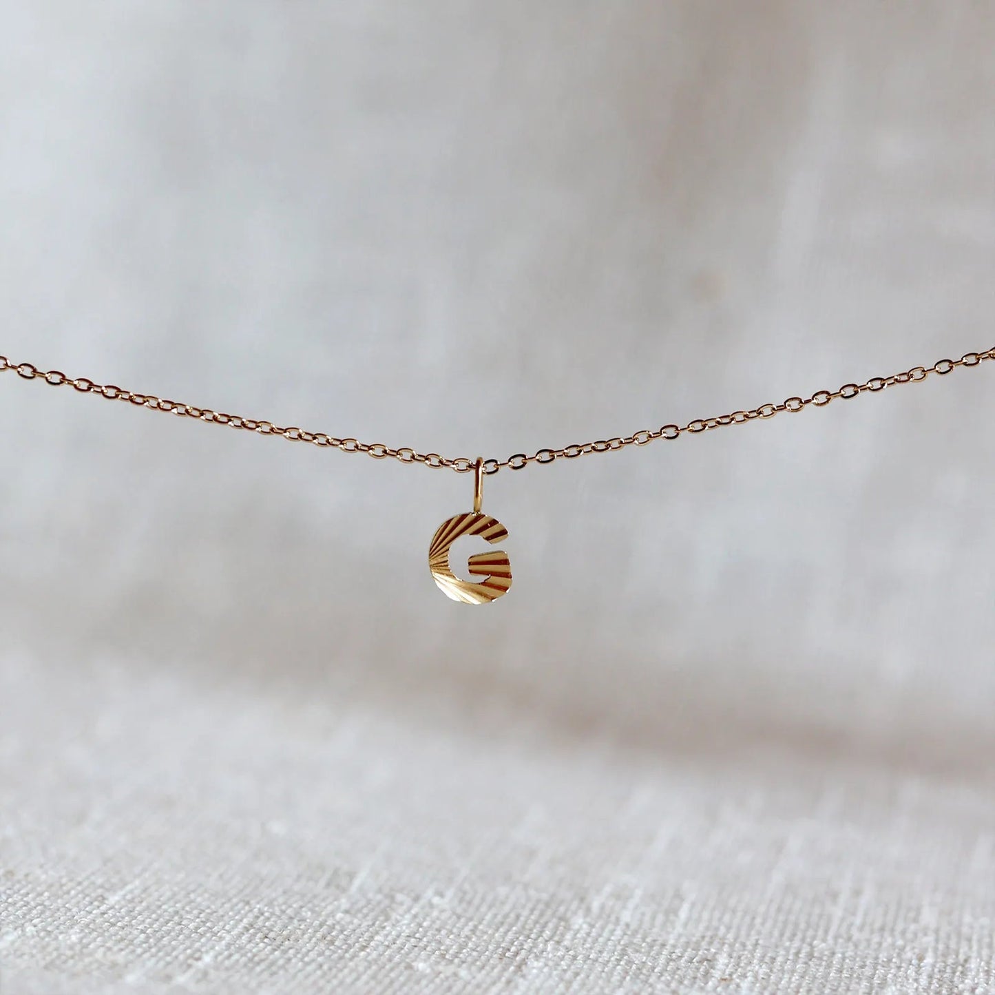 Collier "Je suis" avec pendentif lettre G striée en acier doré, suspendu sur un fond en tissu beige texturé.