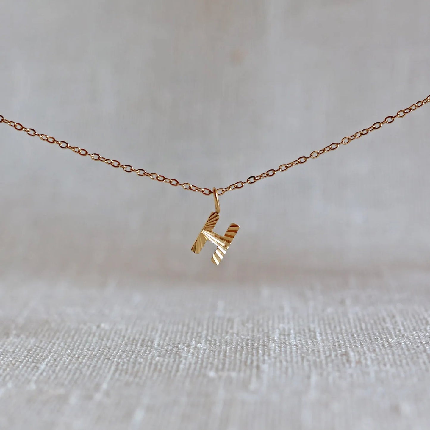 Collier "Je suis" avec pendentif lettre H striée en acier doré, suspendu sur un fond en tissu beige texturé.