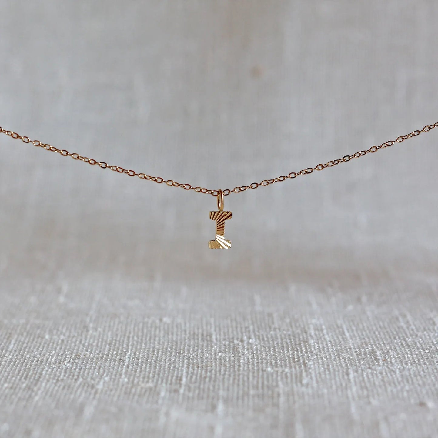 Collier "Je suis" avec pendentif lettre I striée en acier doré, suspendu sur un fond en tissu beige texturé.
