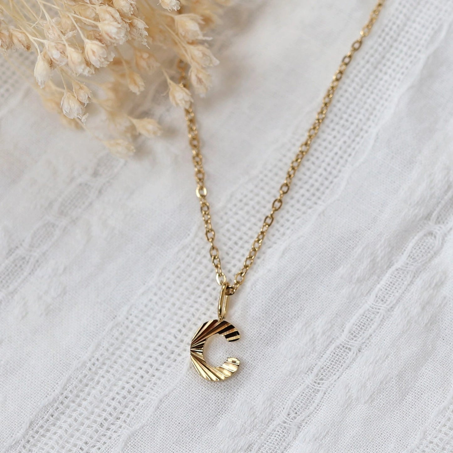 Collier "Je suis" avec pendentif lettre C striée, en acier doré , photographié sur un fond beige texturé.