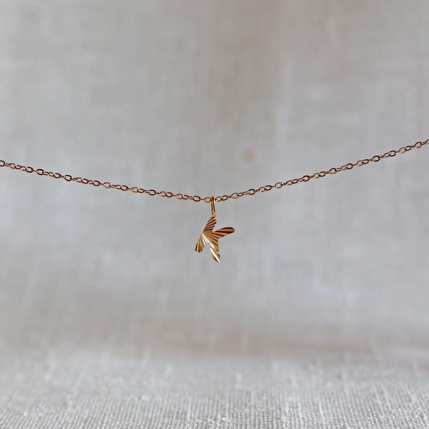 Collier "Je suis" avec pendentif lettre K striée en acier doré, suspendu sur un fond en tissu beige texturé.