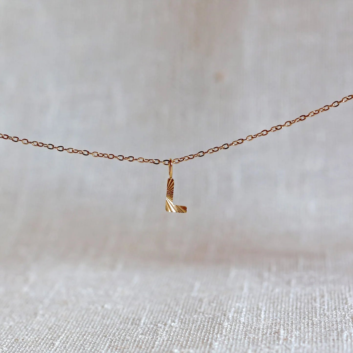 Collier "Je suis" avec pendentif lettre L striée en acier doré, suspendu sur un fond en tissu beige texturé.