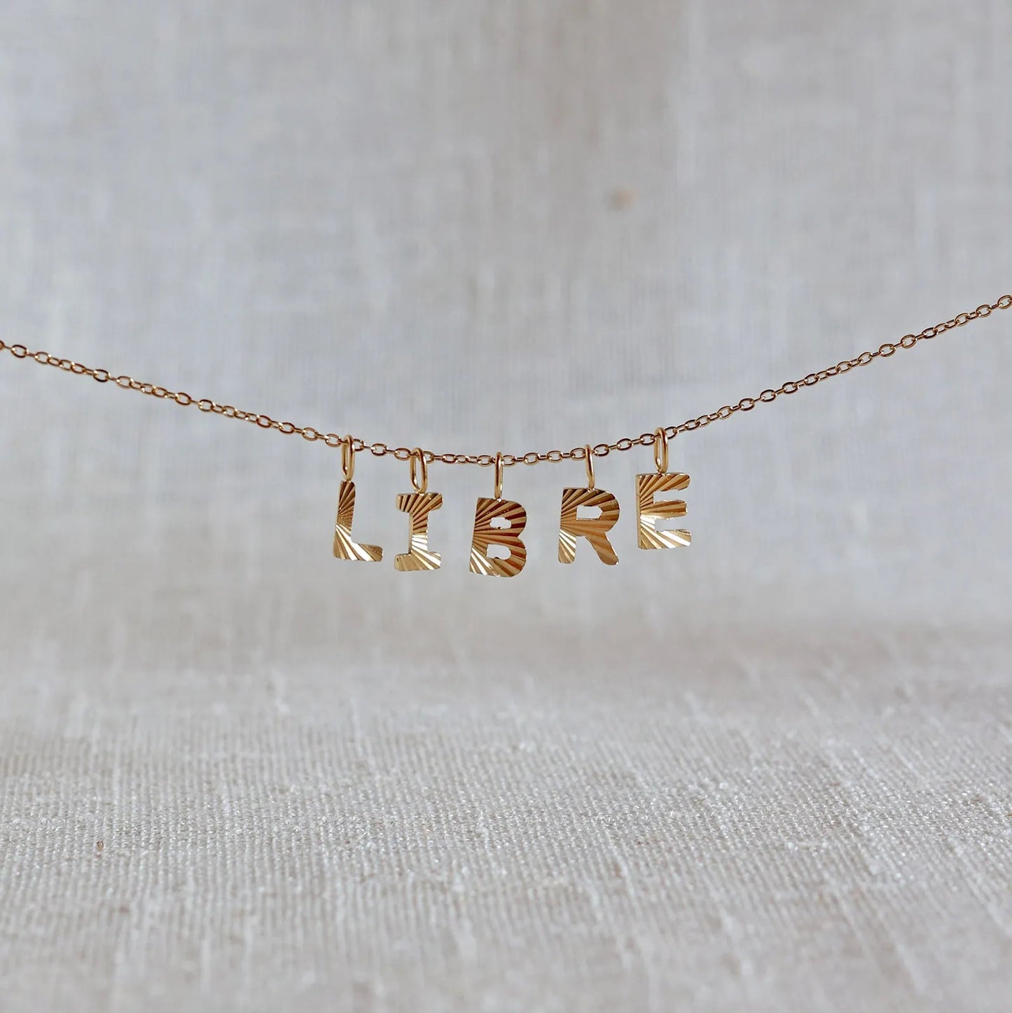Collier "Je suis" avec pendentifs lettres LIBRE striées en acier doré, suspendu sur un fond en tissu beige texturé.