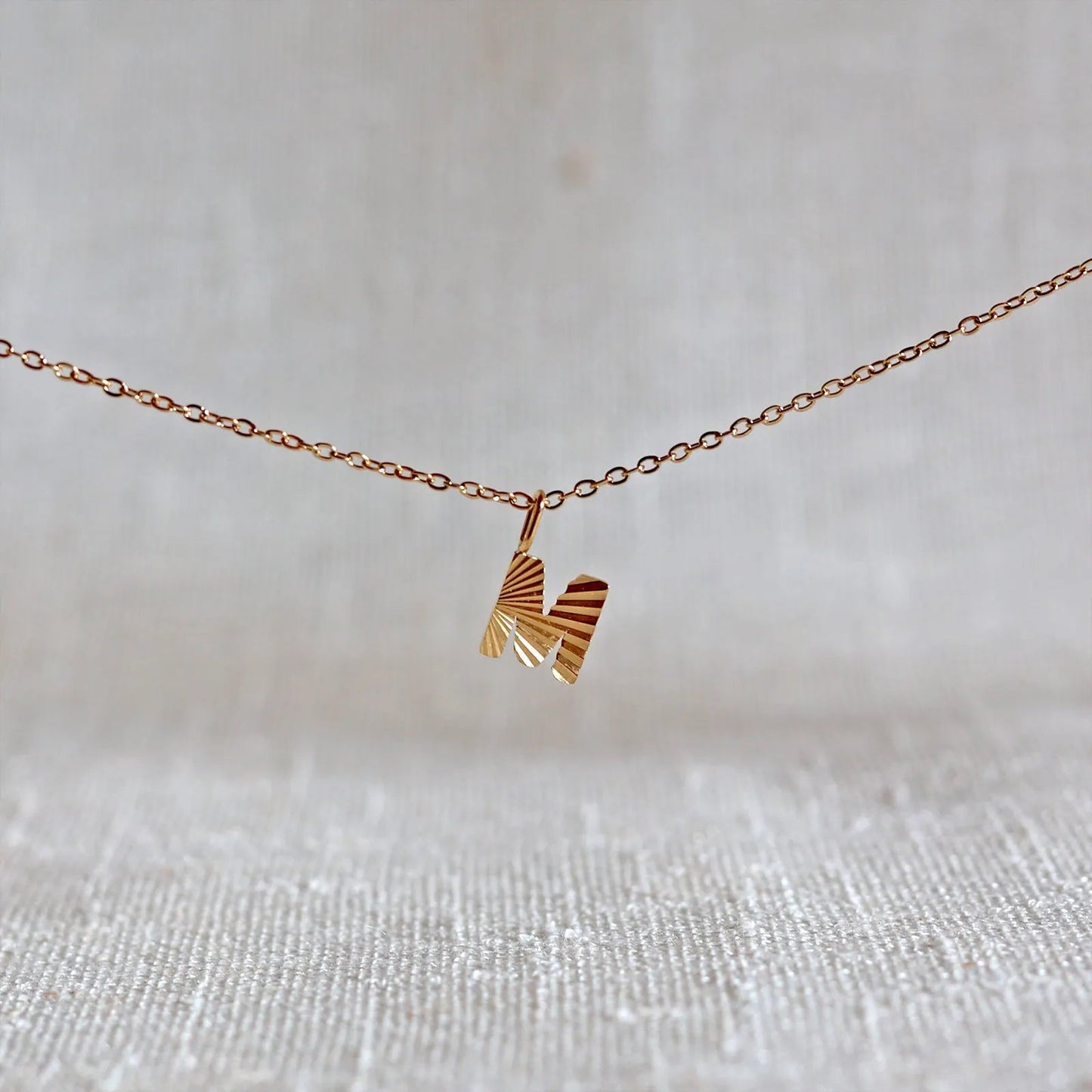 Collier "Je suis" avec pendentif lettre M striée en acier doré, suspendu sur un fond en tissu beige texturé.
