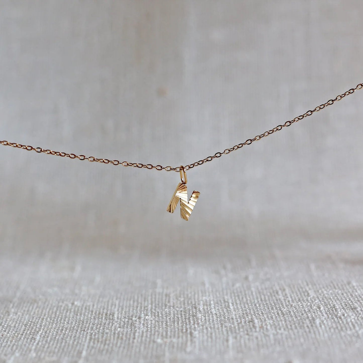 Collier "Je suis" avec pendentif lettre N striée en acier doré, suspendu sur un fond en tissu beige texturé.