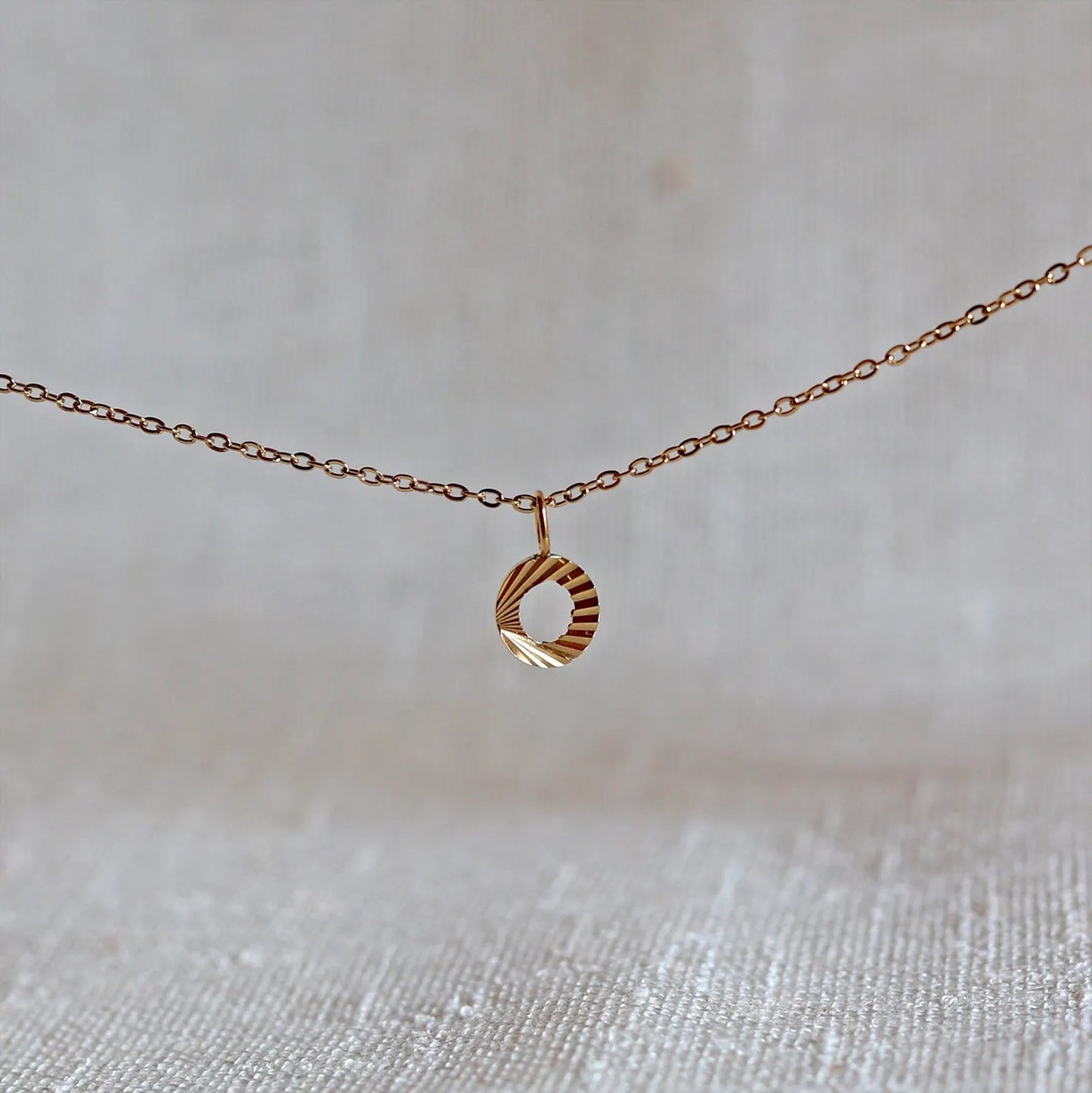 Collier "Je suis" avec pendentif lettre O striée en acier doré, suspendu sur un fond en tissu beige texturé.