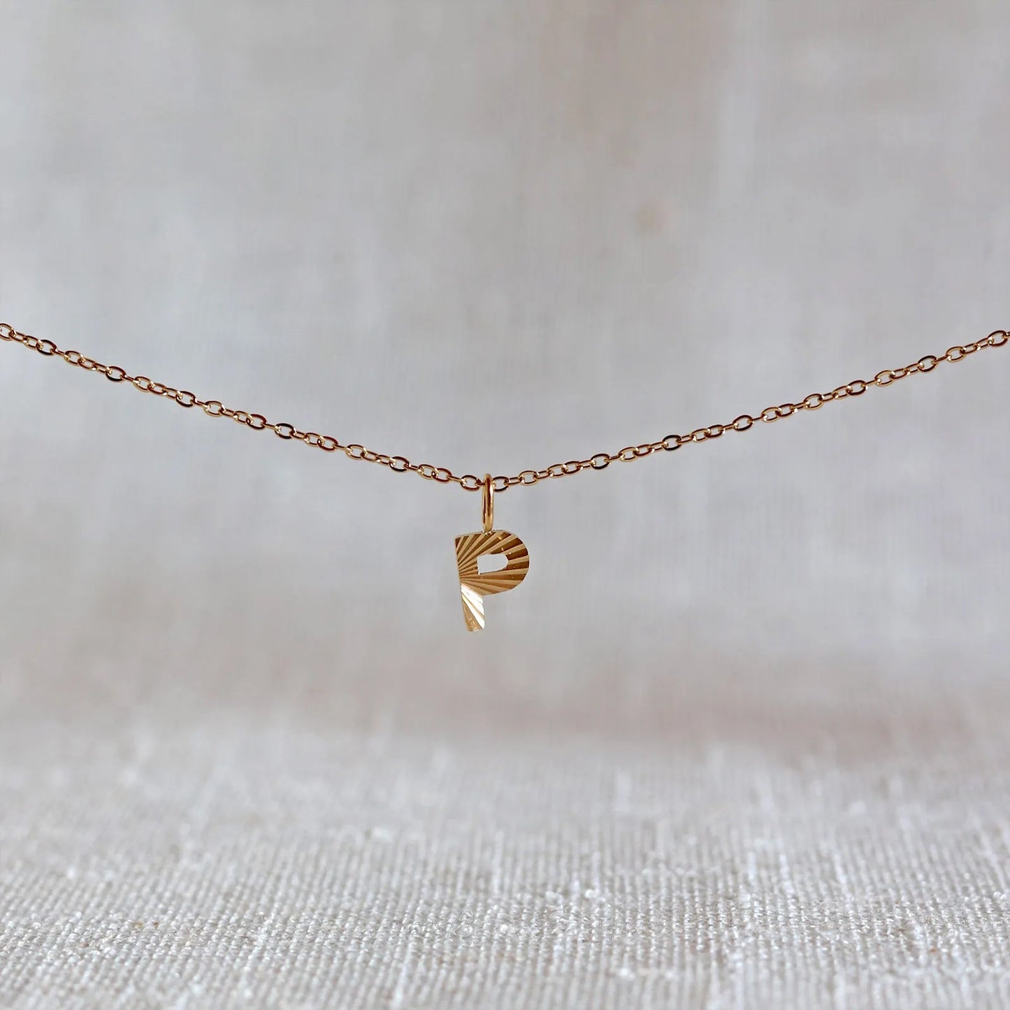 Collier "Je suis" avec pendentif lettre P striée en acier doré, suspendu sur un fond en tissu beige texturé.