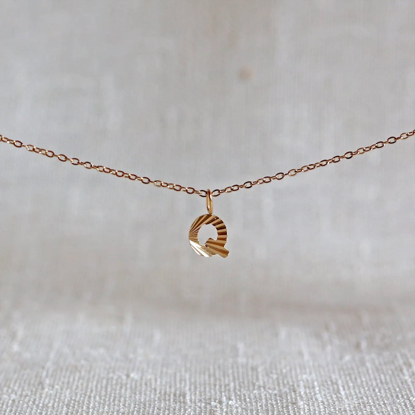 Collier "Je suis" avec pendentif lettre Q striée en acier doré, suspendu sur un fond en tissu beige texturé.