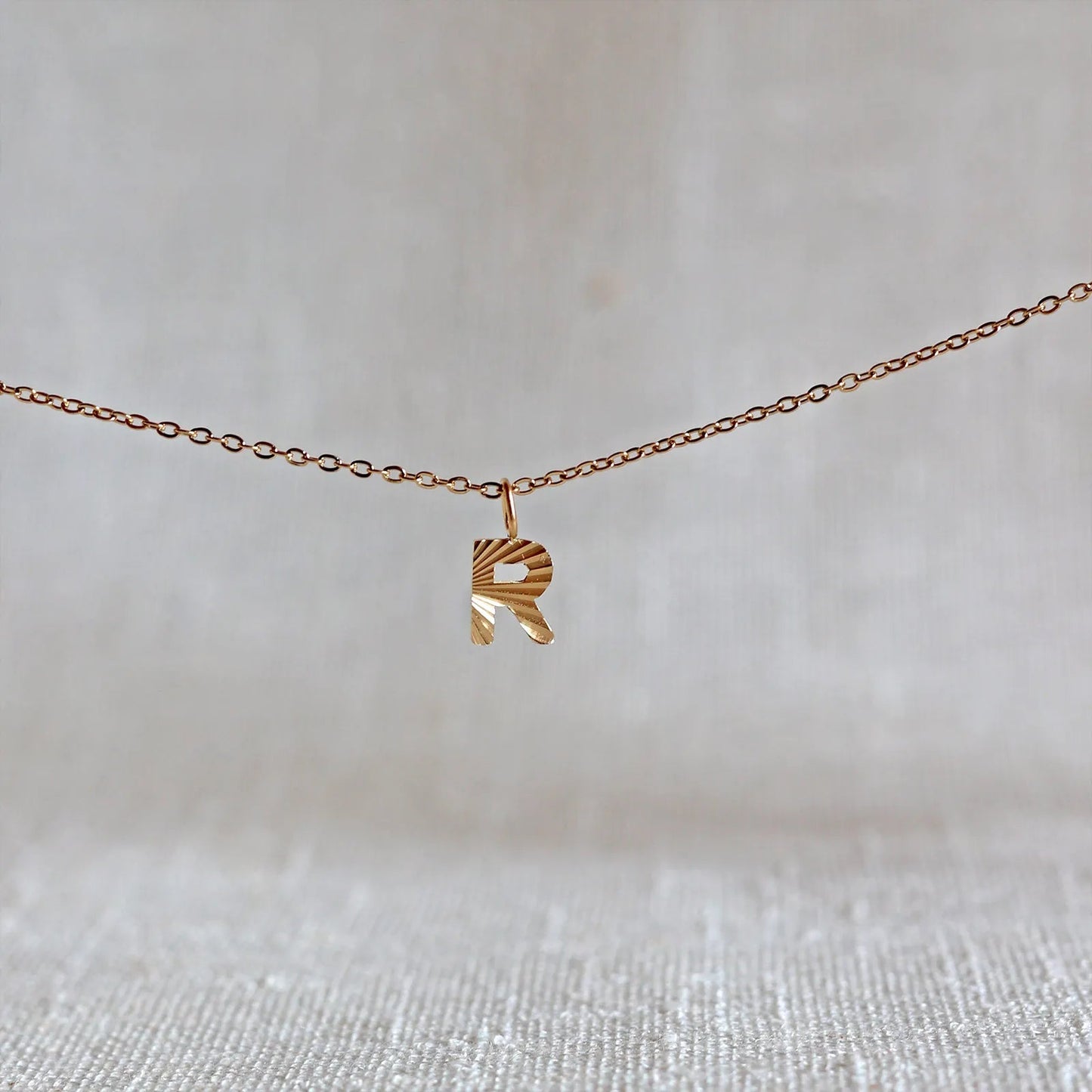 Collier "Je suis" avec pendentif lettre R striée en acier doré, suspendu sur un fond en tissu beige texturé.