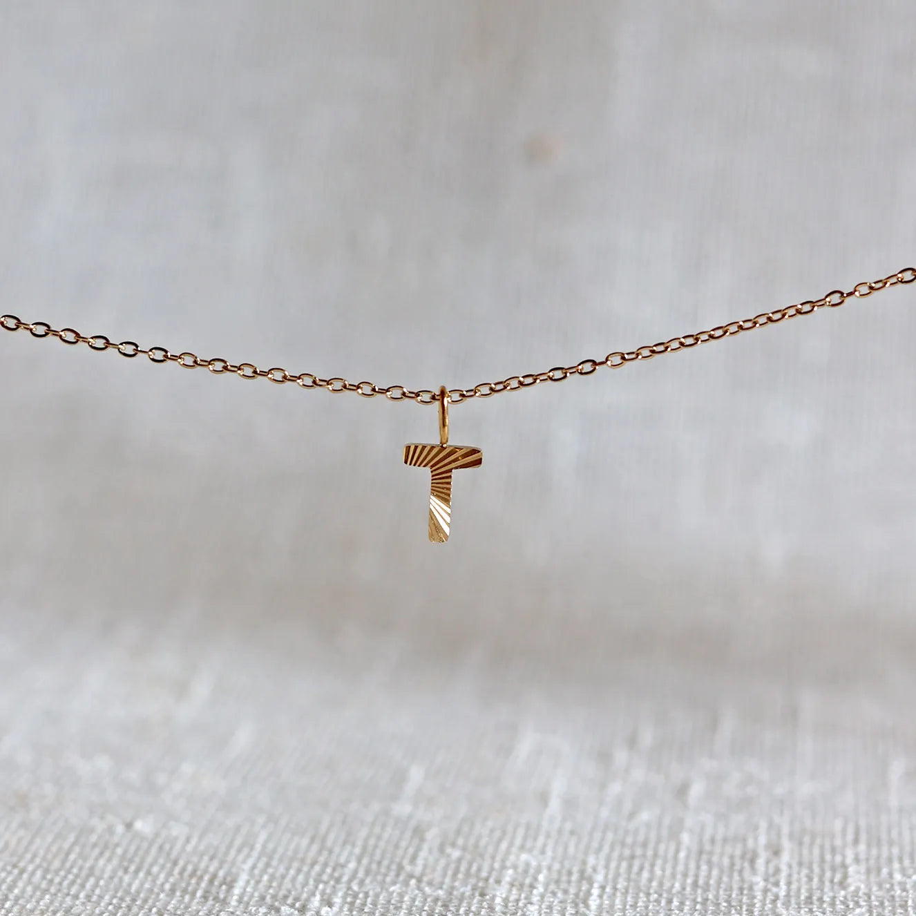 Collier "Je suis" avec pendentif lettre T striée en acier doré, suspendu sur un fond en tissu beige texturé.