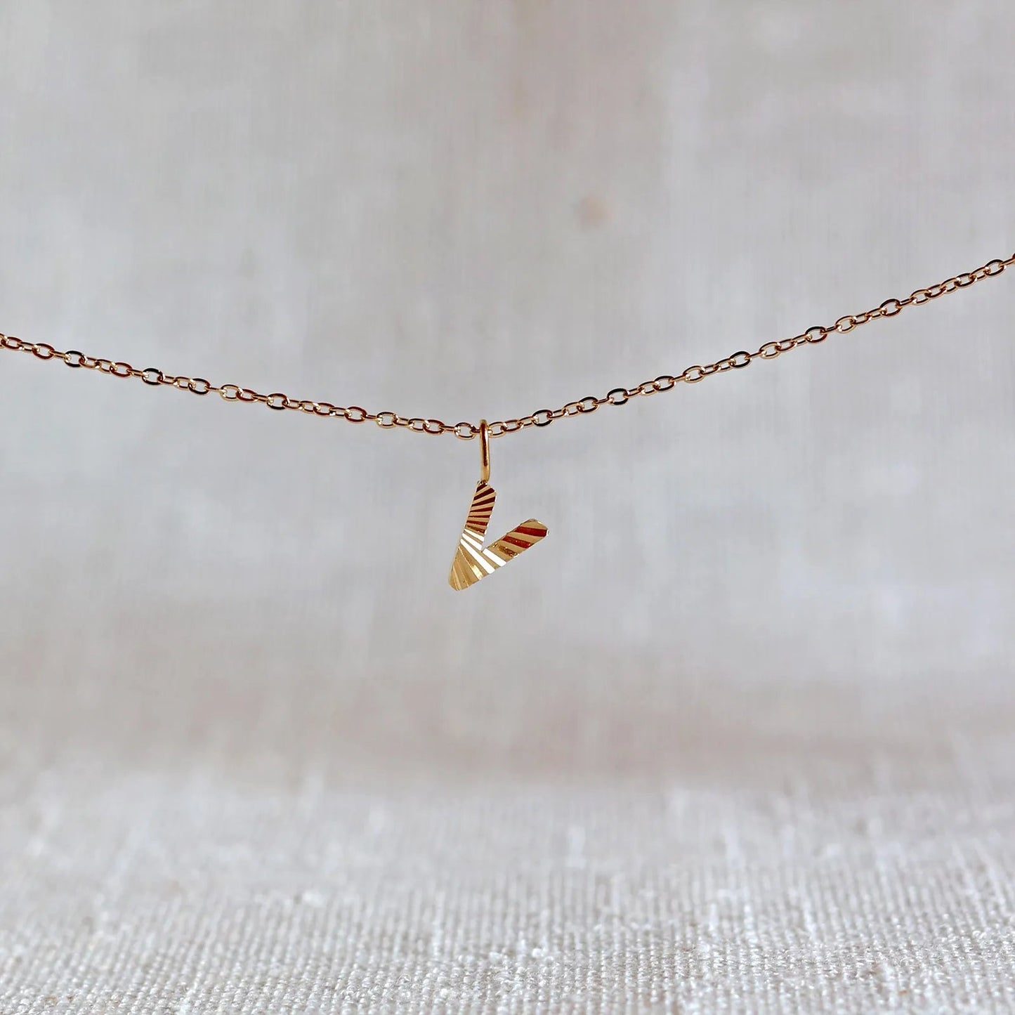 Collier "Je suis" avec pendentif lettre V striée en acier doré, suspendu sur un fond en tissu beige texturé.