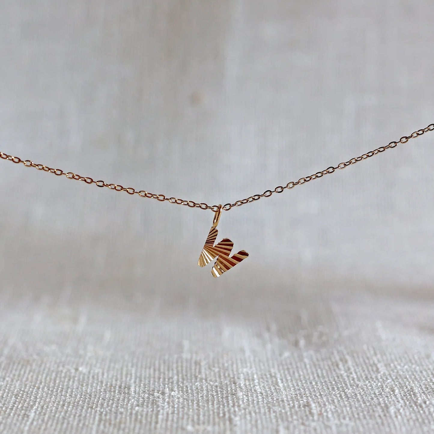 Collier "Je suis" avec pendentif lettre W striée en acier doré, suspendu sur un fond en tissu beige texturé.