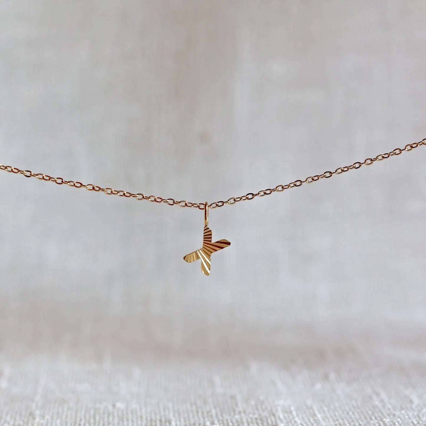 Collier "Je suis" avec pendentif lettre X striée en acier doré, suspendu sur un fond en tissu beige texturé.