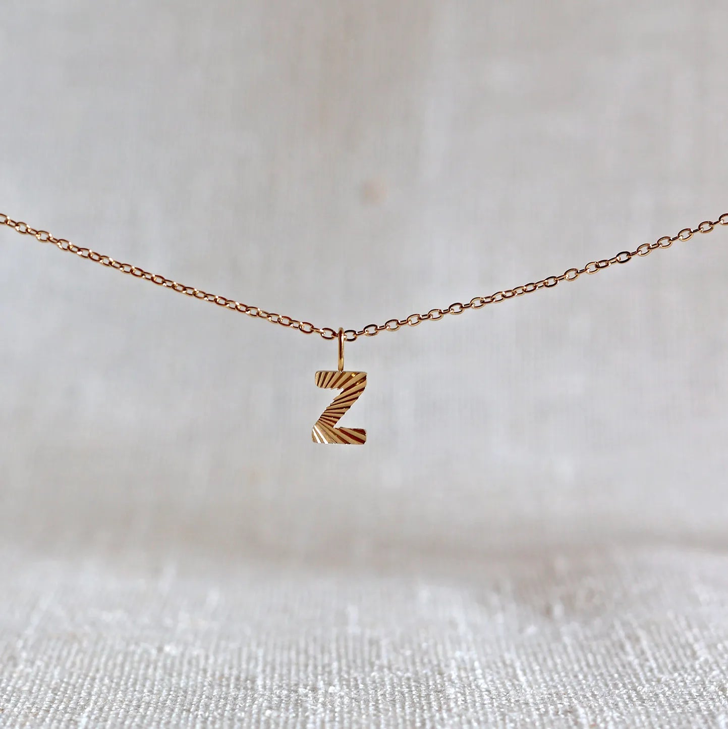 Collier "Je suis" avec pendentif lettre Z striée en acier doré, suspendu sur un fond en tissu beige texturé.