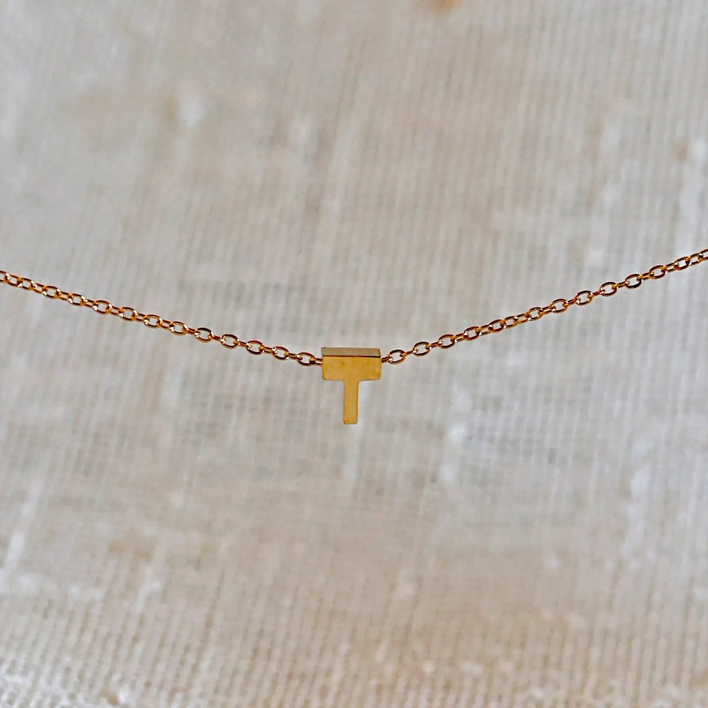 Collier L'instant avec pendentif lettre T doré en acier inoxydable, suspendu au centre d'une maille fine sur fond beige.