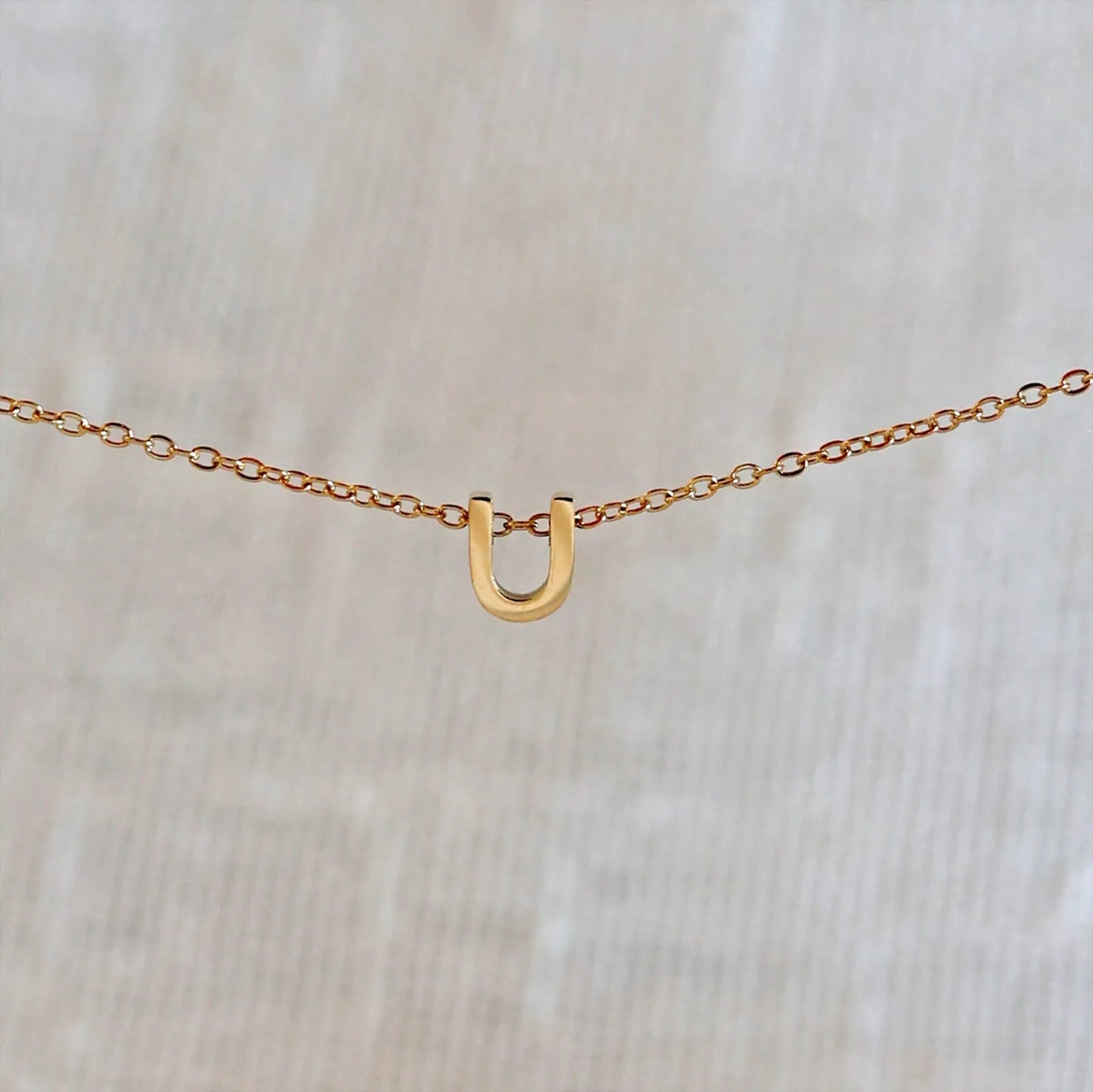 Collier L'instant avec pendentif lettre U doré en acier inoxydable, suspendu au centre d'une maille fine sur fond beige.