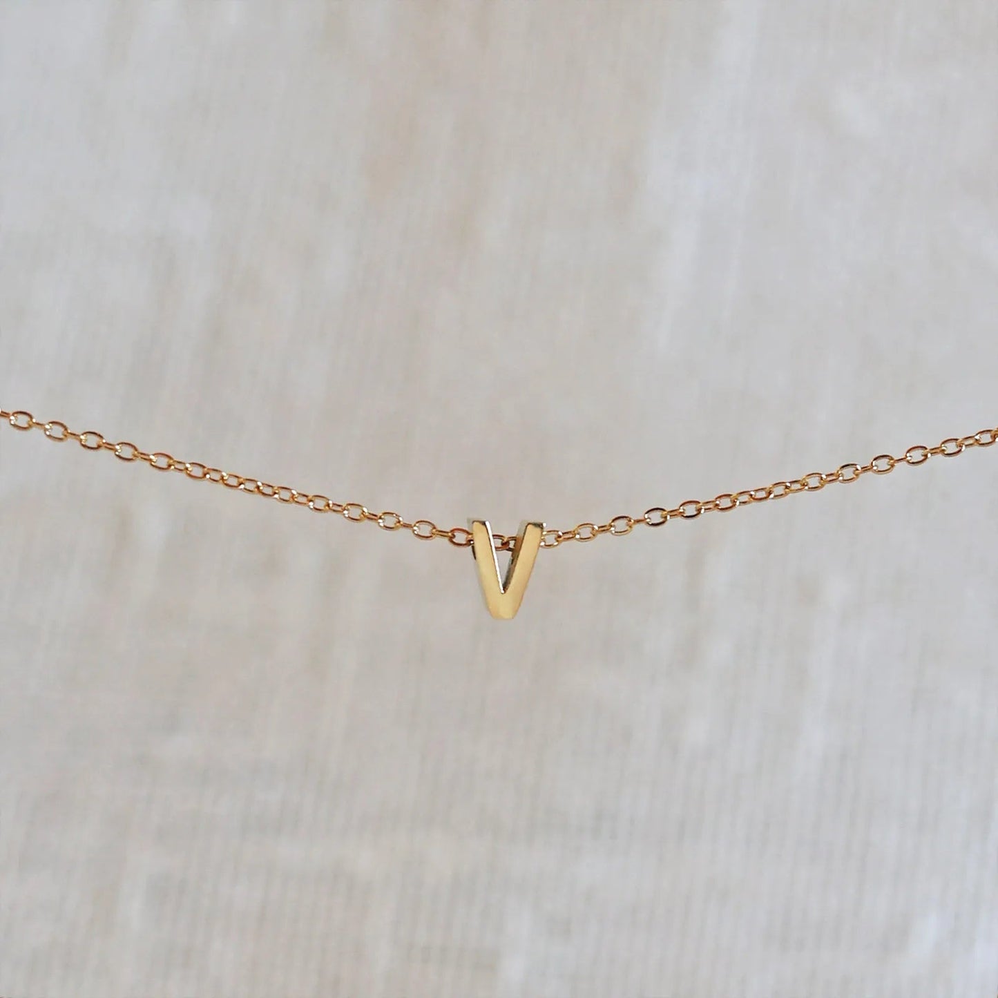 Collier L'instant avec pendentif lettre V doré en acier inoxydable, suspendu au centre d'une maille fine sur fond beige.