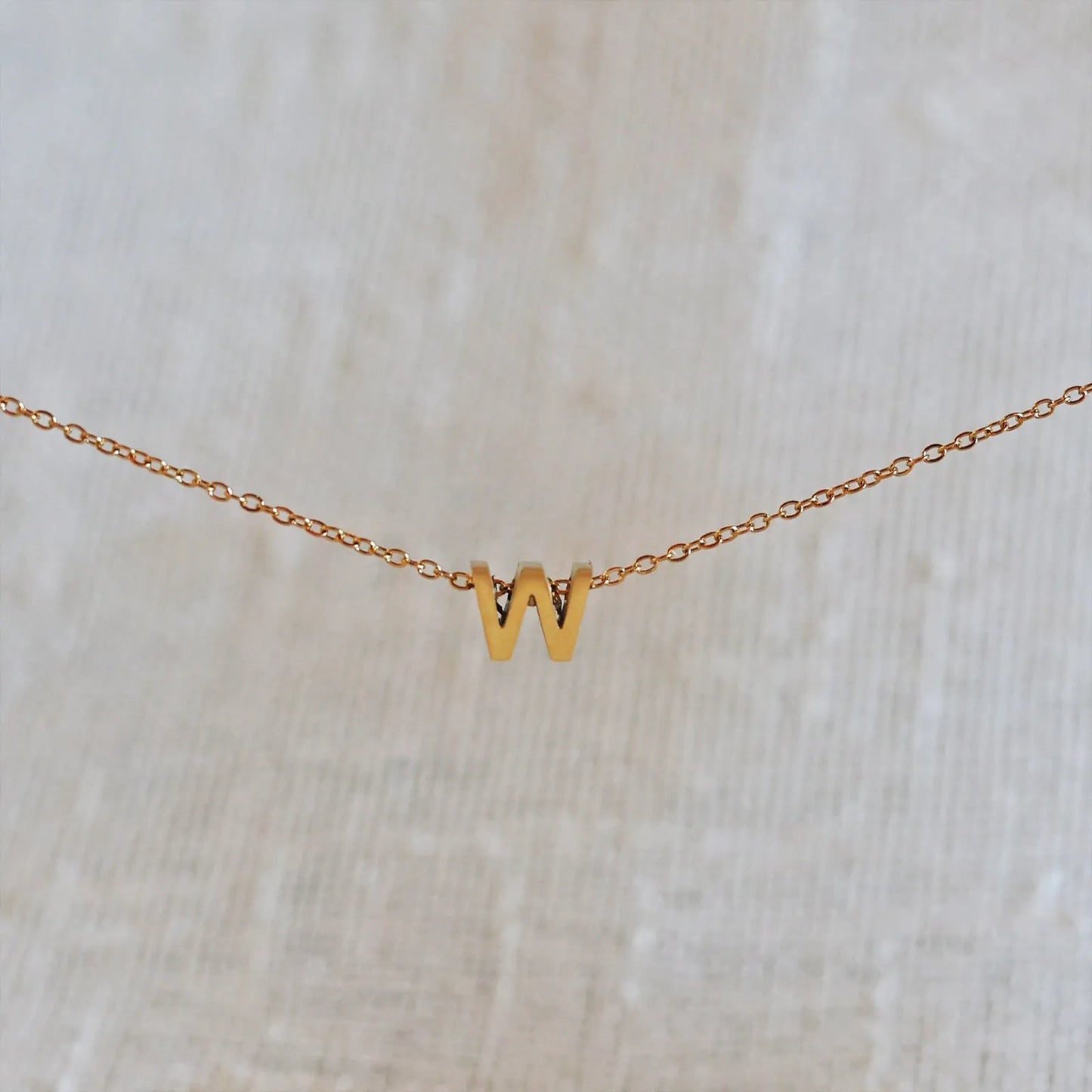 Collier L'instant avec pendentif lettre W doré en acier inoxydable, suspendu au centre d'une maille fine sur fond beige.