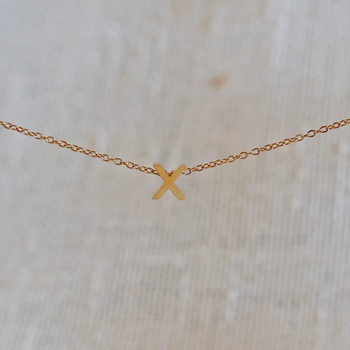 Collier L'instant avec pendentif lettre X doré en acier inoxydable, suspendu au centre d'une maille fine sur fond beige.