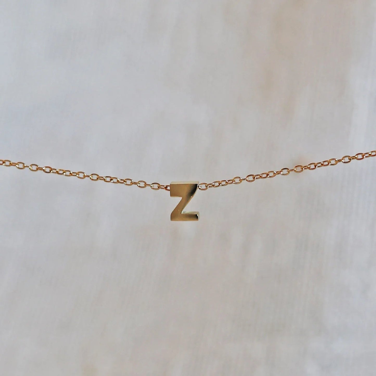 Collier L'instant avec pendentif lettre Z doré en acier inoxydable, suspendu au centre d'une maille fine sur fond beige.