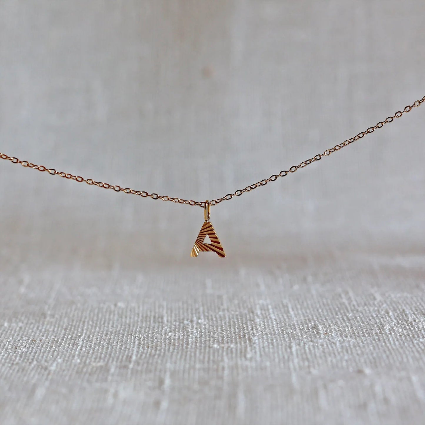 Collier "Je suis" avec pendentif lettre A striée en acier doré, suspendu sur un fond en tissu beige texturé.