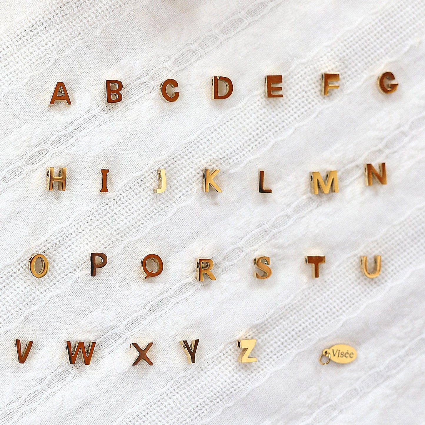 Ensemble de pendentifs lettres dorées en acier inoxydable, soigneusement alignés sur un tissu beige texturé.