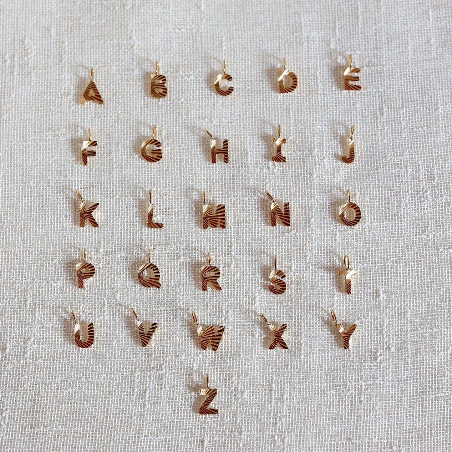 Ensemble de pendentifs lettres dorées en acier inoxydable, soigneusement alignés sur un tissu beige texturé.