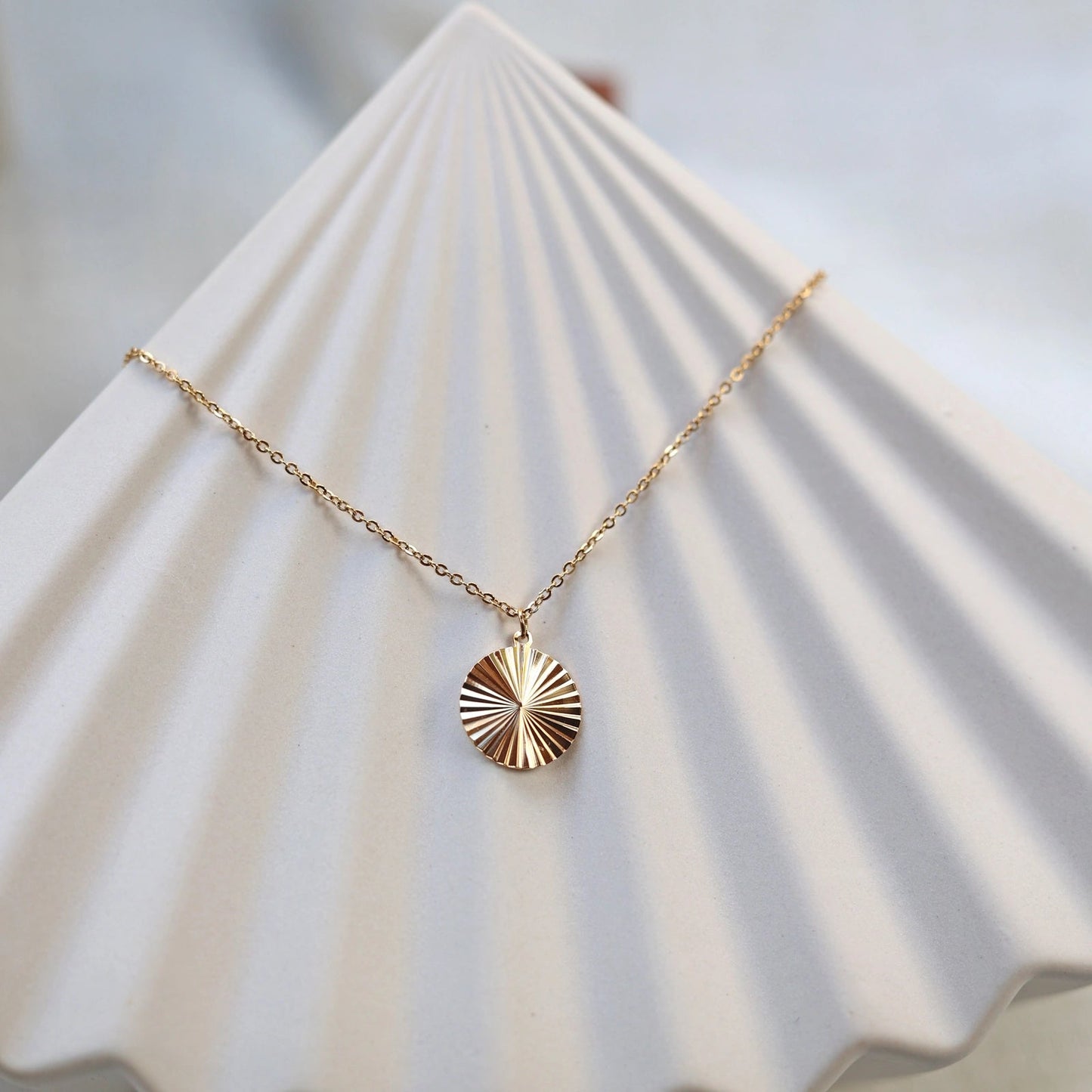 Vue de face du collier "Energie", pendentif rond strié personnalisable en acier doré, posé sur une structure beige.