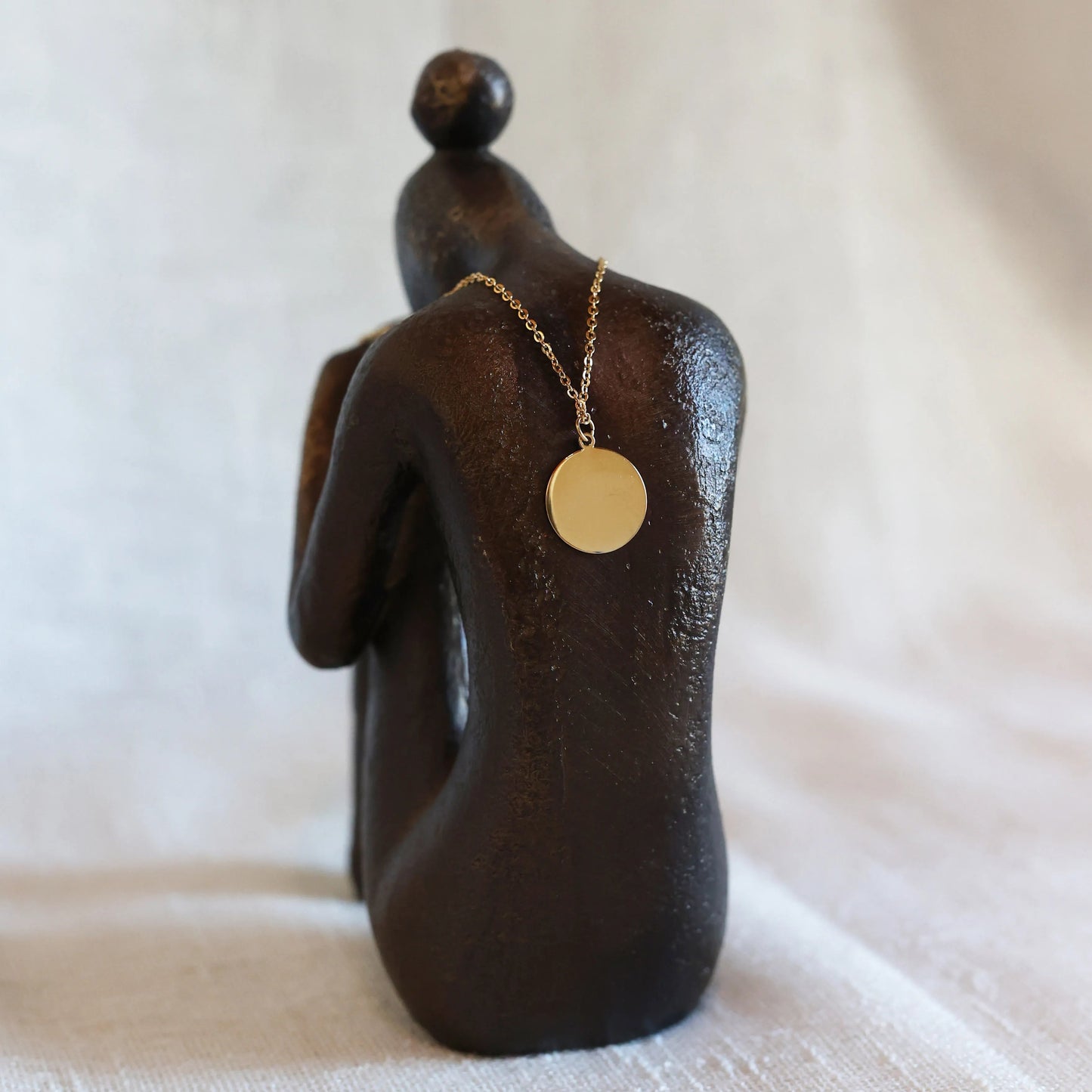Vue arrière du collier "Energie", pendentif rond strié personnalisable en acier doré, posé sur une statue de femme de dos.