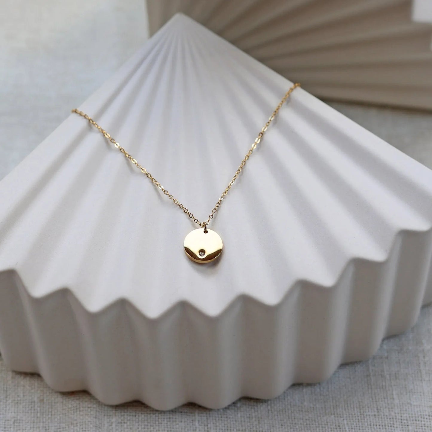Collier personnalisable "Lumineuse", orné d'une médaille ronde brillante en acier doré, posé sur un fond beige texturé.