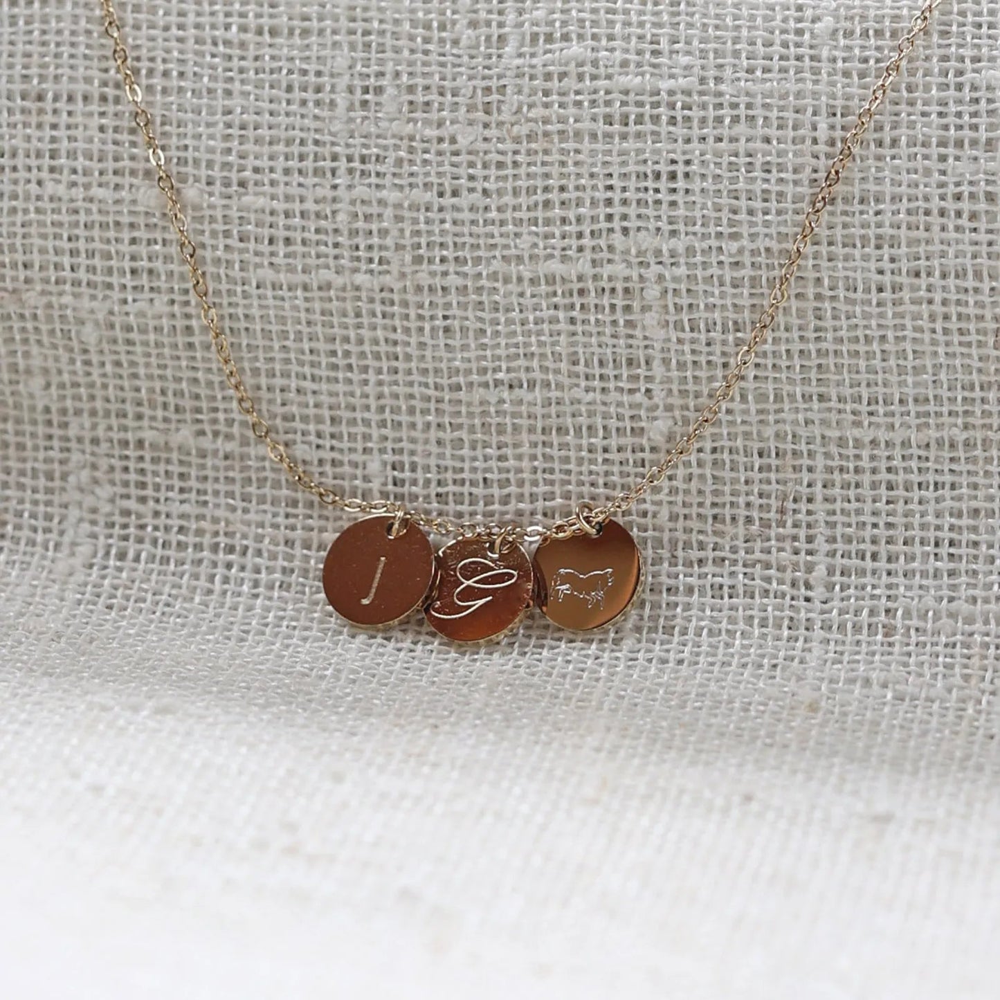 Collier "Inspiration" en acier inoxydable doré, composé de trois médaillons personnalisés d'initiales et dessin, présenté sur un fond beige texturé.