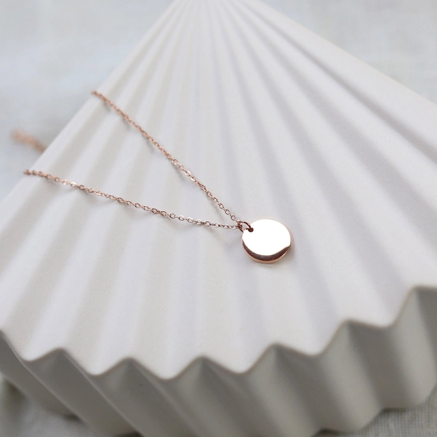 Collier personnalisable "Lumineuse", orné d'une médaille ronde brillante en acier or rose, posé sur un fond beige texturé.