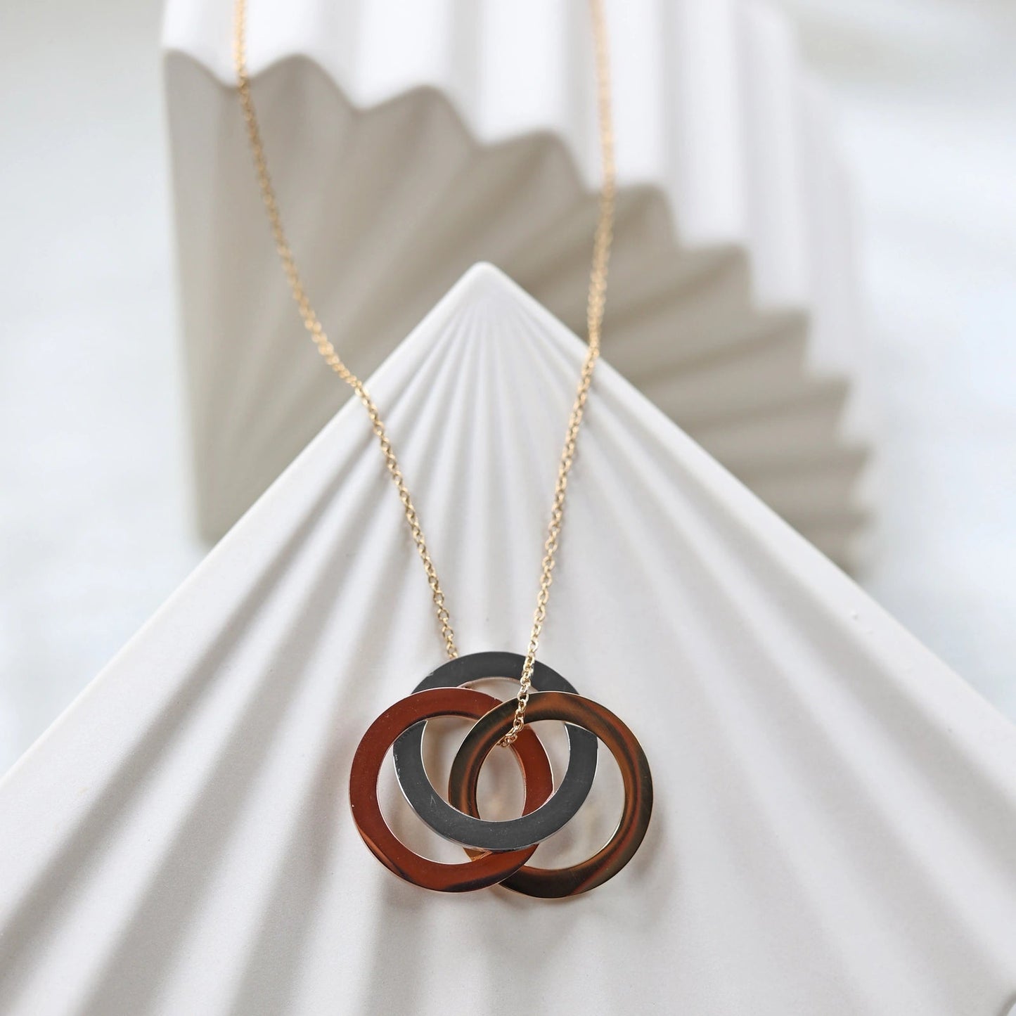 Collier "Je deviens" en acier argenté, avec trois cercles entrelacés personnalisables, posé sur un fond beige texturé.